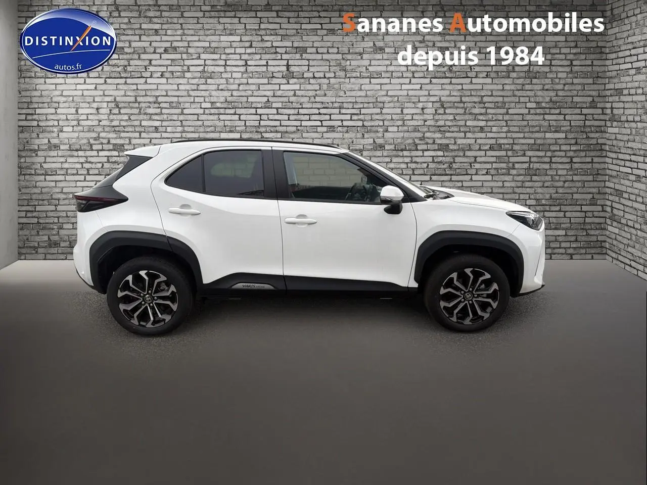 Profil côté gauche de la Toyota Yaris Cross Hybrid 130h blanche avec jantes alliage et protections noires sur fond de mur en briques.