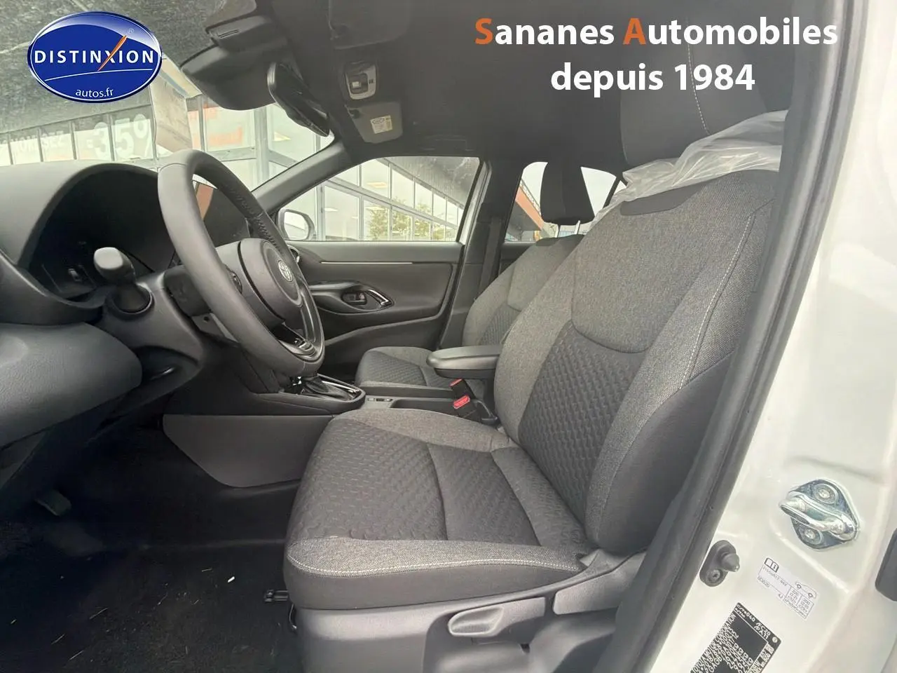 Vue intérieure côté conducteur de la Toyota Yaris Cross Hybrid blanche 2025, sièges tissu gris et volant multifonction.