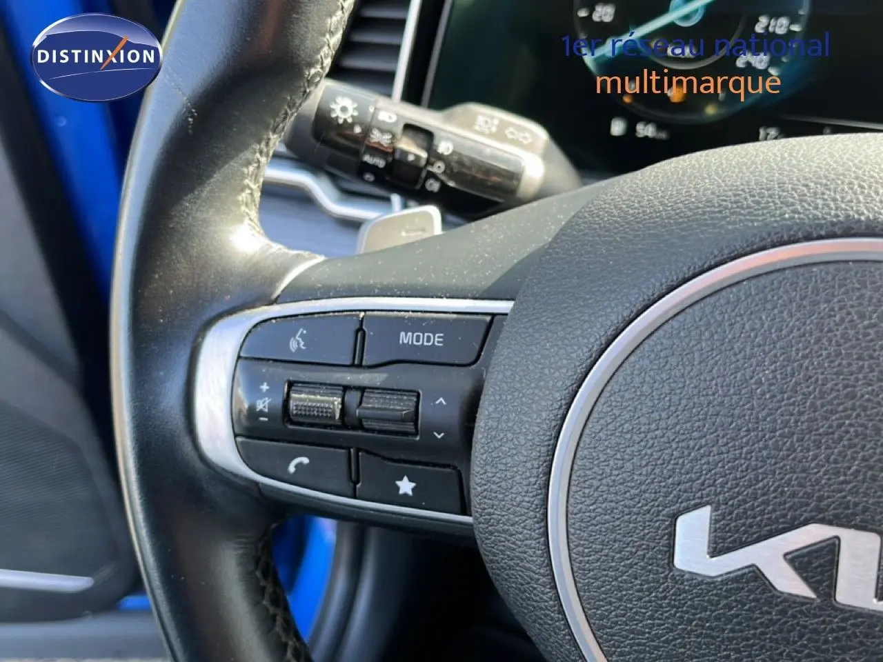 Gros plan sur le volant multifonction noir du Kia Sportage bleu fusion métal, avec commandes audio et tableau de bord en arrière-plan.