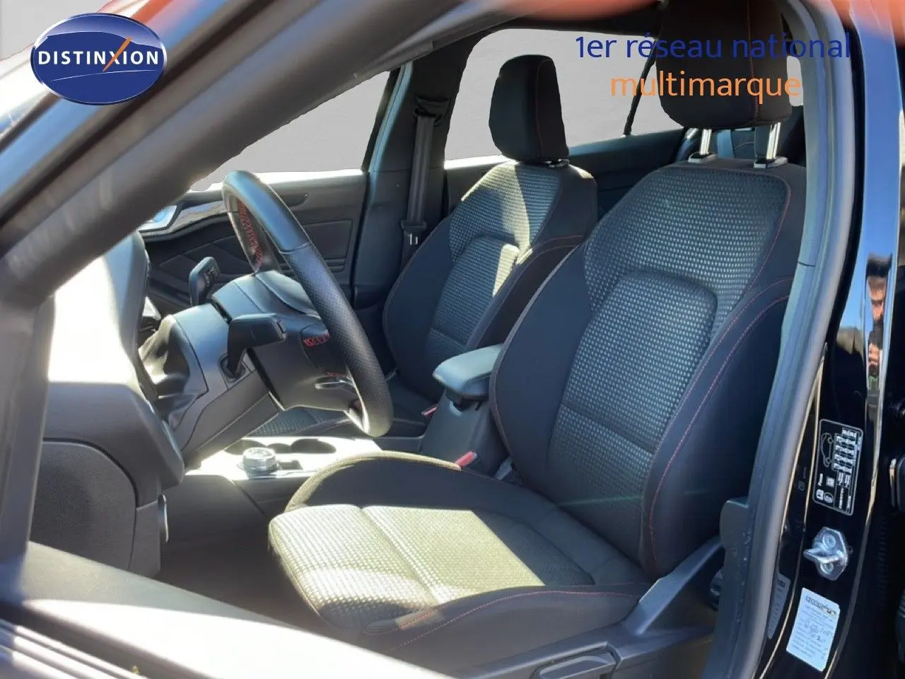 Vue intérieure côté conducteur de la Ford Focus SW noire, sièges tissu avec surpiqûres rouges et volant cuir multifonction.