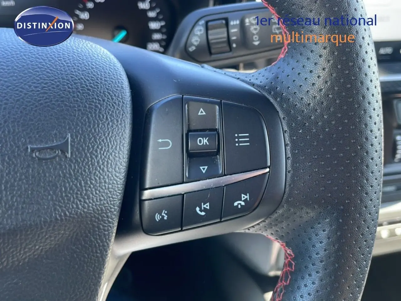 Gros plan sur le volant en cuir noir perforé avec surpiqûres rouges de la Ford Focus SW 2024, boutons de commande visibles.