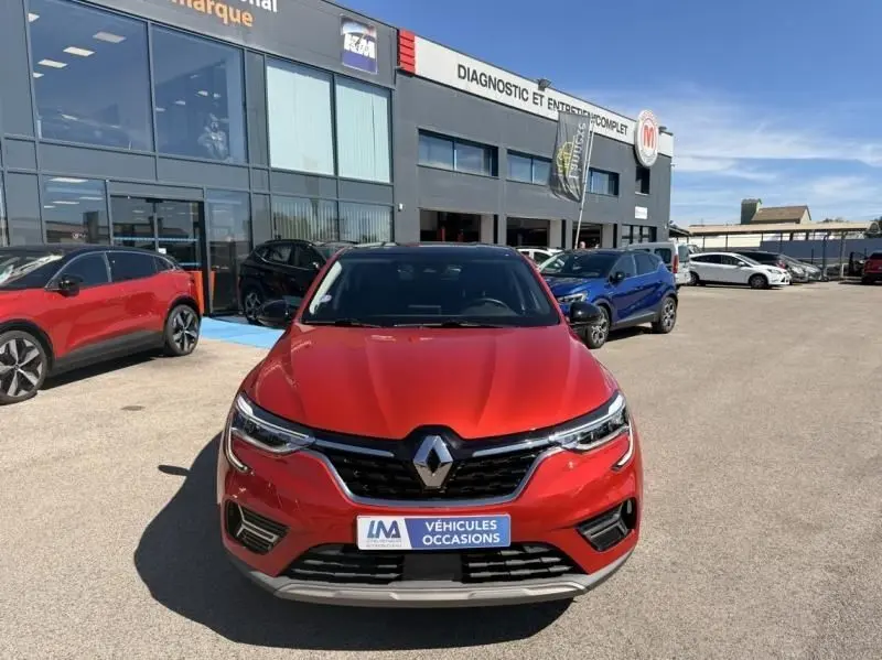 Vue avant d'un Renault Arkana rouge flamme 2022 avec calandre noire et logo chromé, stationné devant un garage.
