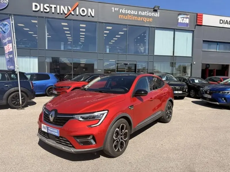 Renault Arkana rouge flamme en 3/4 avant droit, avec jantes distinctives et toit noir, stationné devant un showroom multimarque.