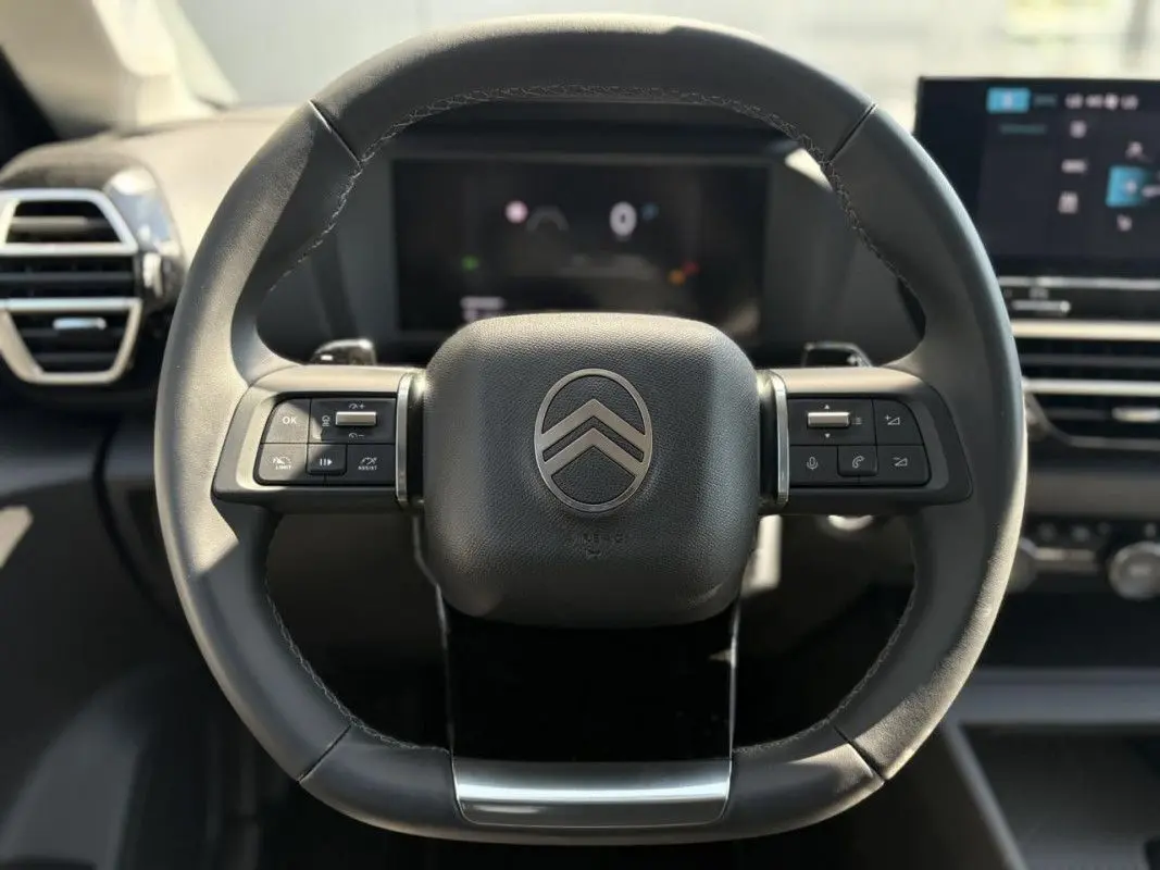 Gros plan sur le volant noir du Citroën C4 Hybrid 145 ch, avec commandes intégrées et tableau de bord numérique en arrière-plan.