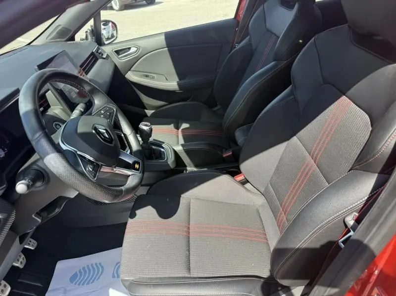 Vue intérieure côté conducteur de la Renault Clio V rouge flamme 2022, sièges sport noirs avec surpiqûres rouges et boîte manuelle.