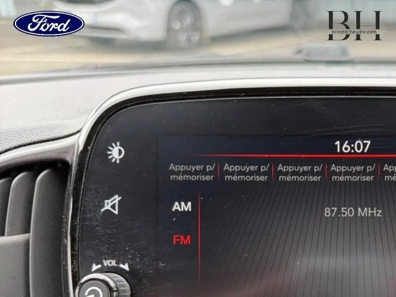 Tableau de bord d'une Ford vu de près, écran tactile affichant la radio FM à 87,50 MHz, volant partiellement visible.