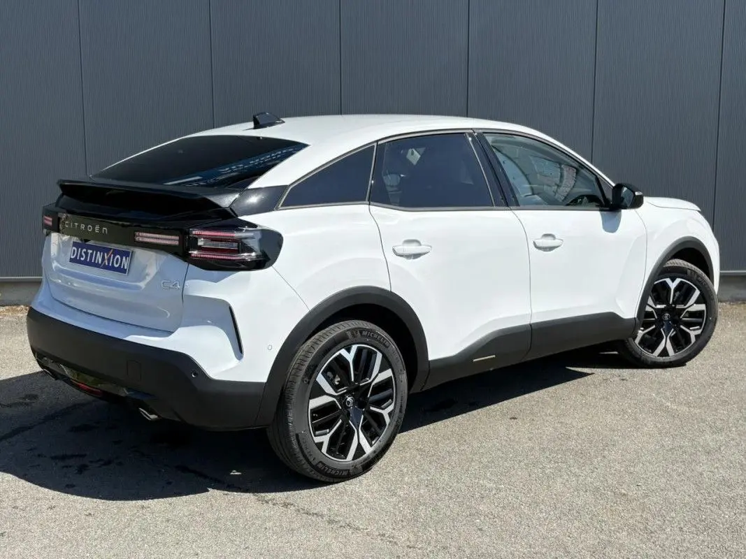 Vue de profil côté gauche d'une Citroën C4 Hybrid blanche Okenite avec jantes noires et toit noir, stationnée devant un mur métallique.