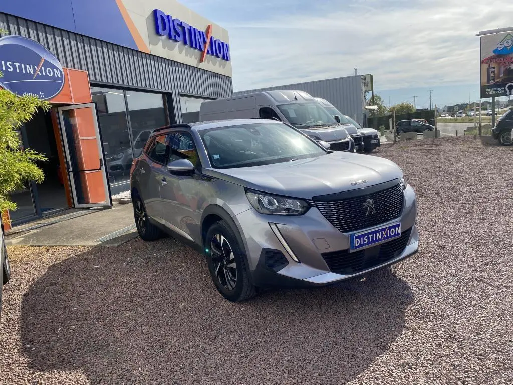 Peugeot 2008 gris clair vue 3/4 avant droit, avec calandre noire et jantes alliage noires, devant un bâtiment Distinxion.