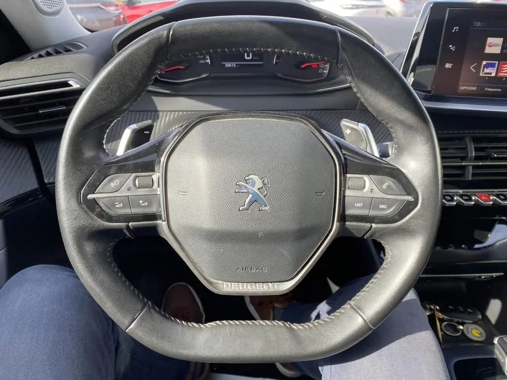Vue rapprochée du volant cuir noir du Peugeot 2008 2020 avec commandes intégrées et tableau de bord numérique en arrière-plan.