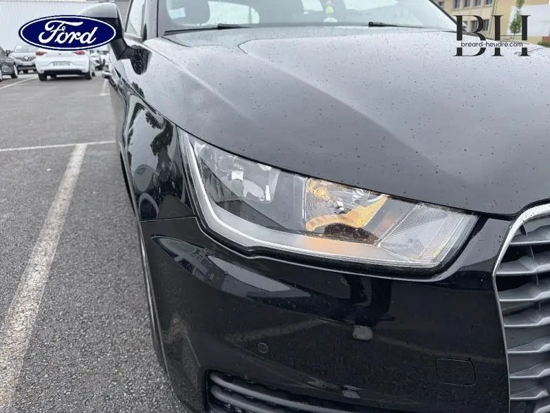Gros plan sur le phare avant gauche d'une Audi A1 Sportback noire brillante sous la pluie, avec calandre partielle visible.