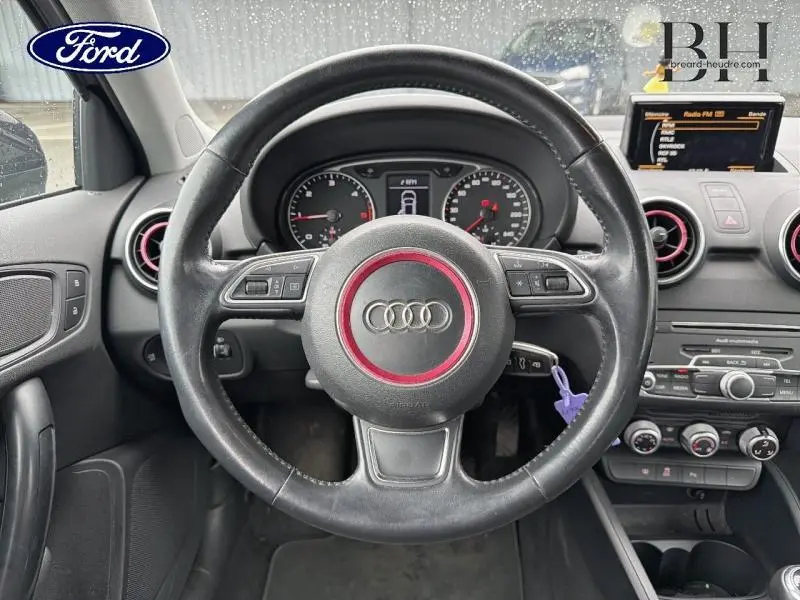 Vue intérieure centrée sur le volant cuir noir avec logo Audi rouge, tableau de bord et écran multifonction du A1 Sportback.