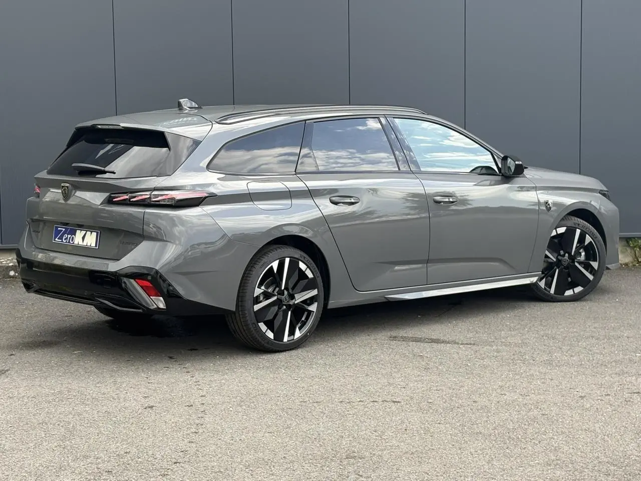 Peugeot 308 SW gris Sélénium vue 3/4 arrière droit, jantes alliage bicolores et barres de toit noires.