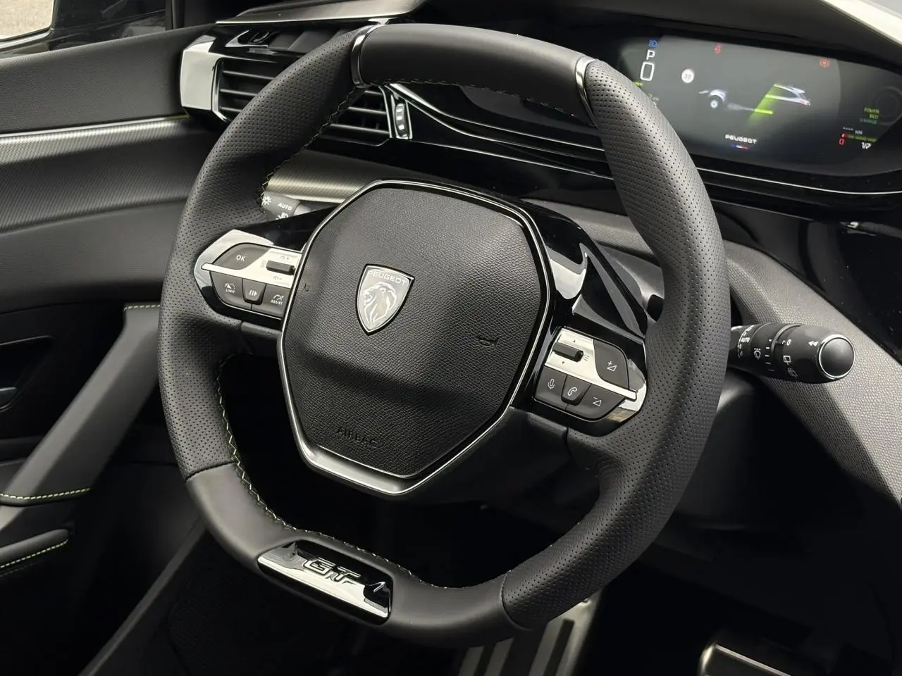 Vue rapprochée du volant GT noir avec surpiqûres vert Adamite et tableau de bord numérique du Peugeot 308 SW Hybrid 2026.