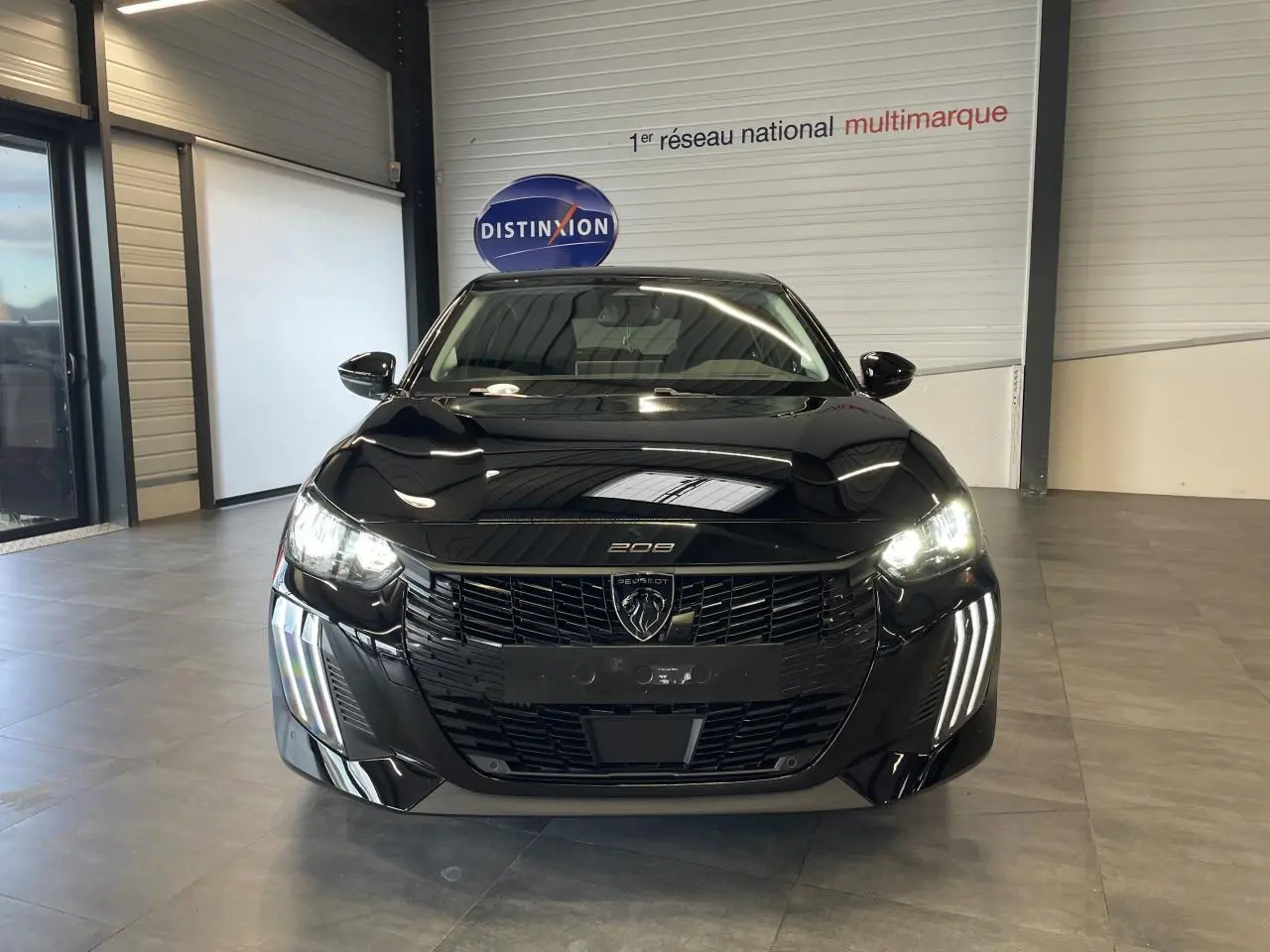 Vue avant d'une Peugeot 208 noire 2025 avec phares allumés et calandre noire brillante en intérieur showroom.