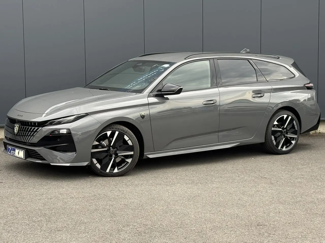Peugeot 308 SW gris Sélénium vue 3/4 avant droit avec jantes bi-tons et barres de toit noires.