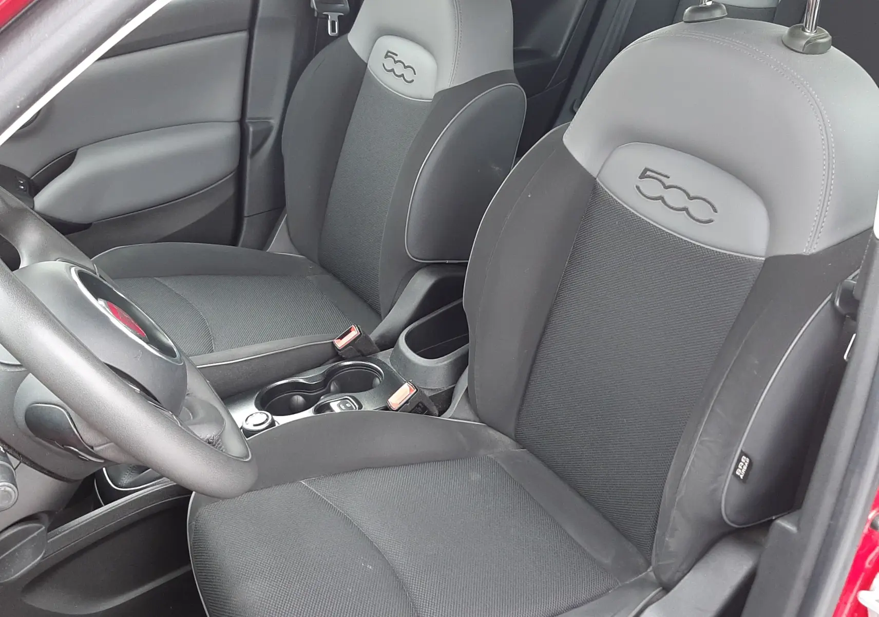 Vue intérieure côté conducteur d'une FIAT 500X rouge, sièges gris foncé avec logo 500 embossé sur les appuie-têtes.