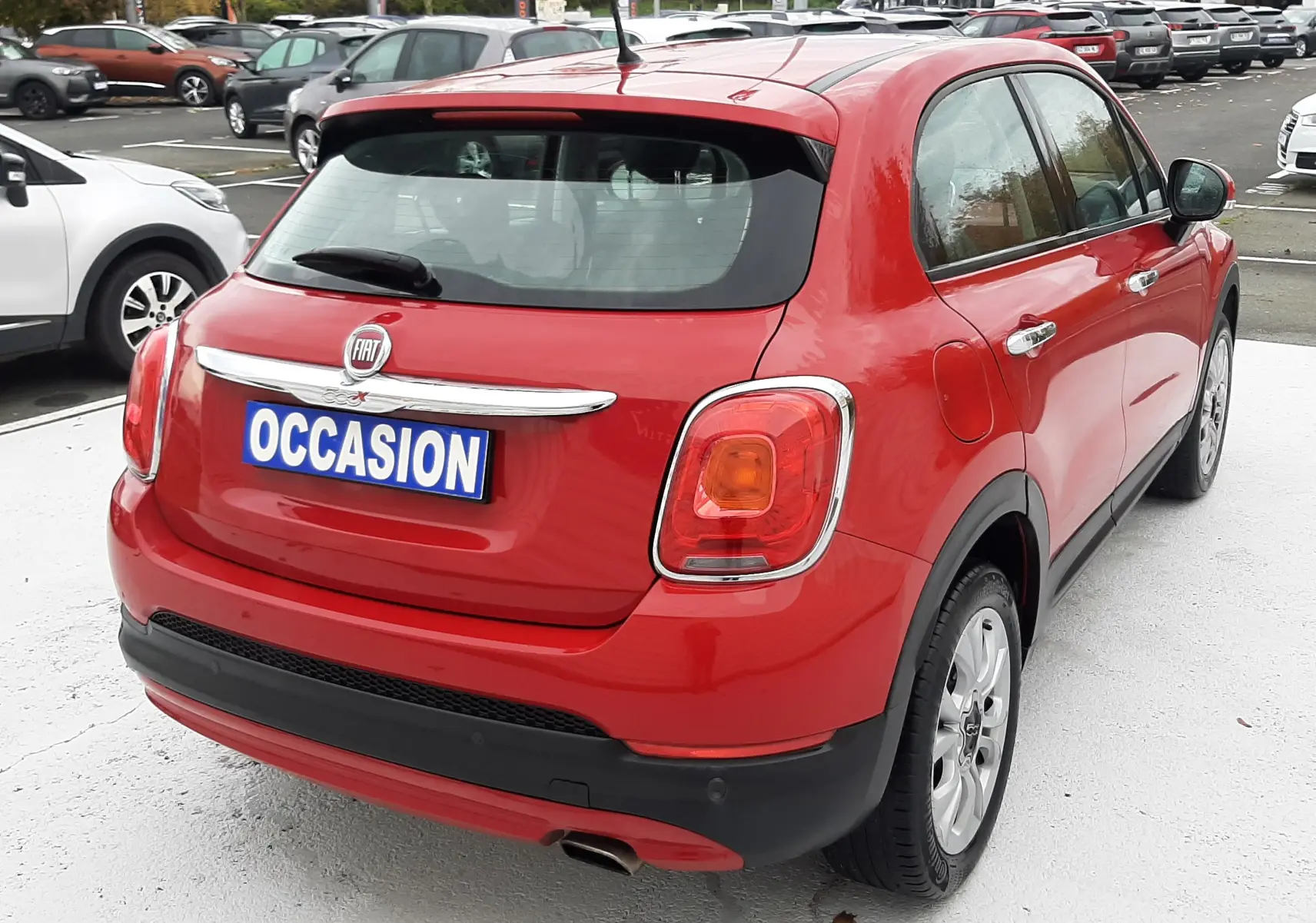 Vue 3/4 arrière droite d'une FIAT 500X rouge avec plaque "OCCASION" sur parking extérieur.