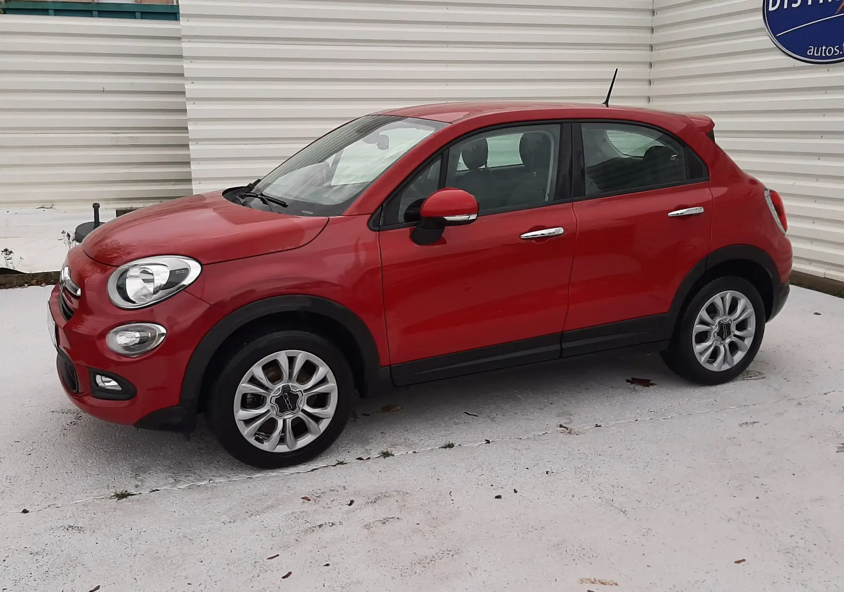Vue de profil côté gauche d'une Fiat 500X rouge 5 portes garée devant un mur blanc avec logo bleu.