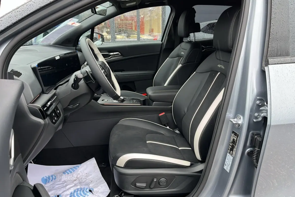 Vue intérieure côté conducteur de la KIA Sportage 2026 gris perle, sièges sport noirs avec surpiqûres blanches et volant cuir multifonctions.
