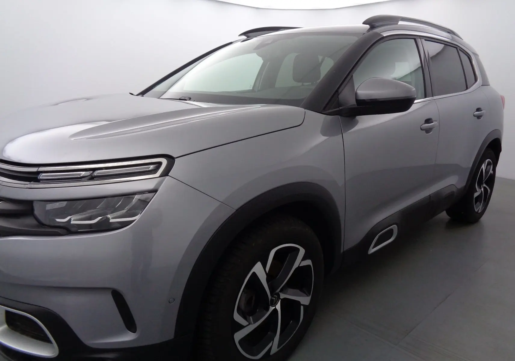 Vue 3/4 avant droite d'un Citroën C5 Aircross gris Artense métal avec jantes bi-ton et barres de toit noires.