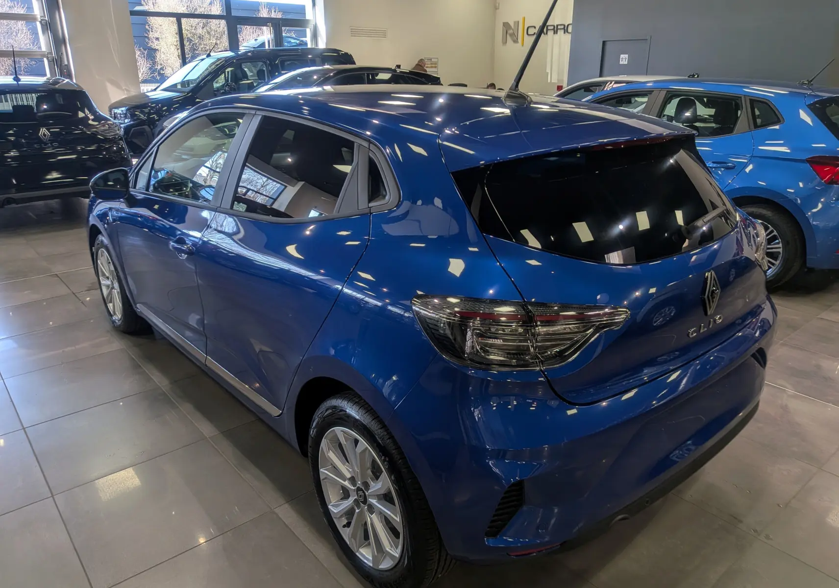 Renault Clio V Bleu Iron vue 3/4 arrière droit en intérieur, avec feux arrière fumés et jantes alliage.