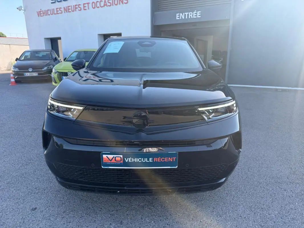 Vue avant d’un Opel Mokka électrique noir 2025, avec calandre lisse et phares LED fins, stationné devant un garage.