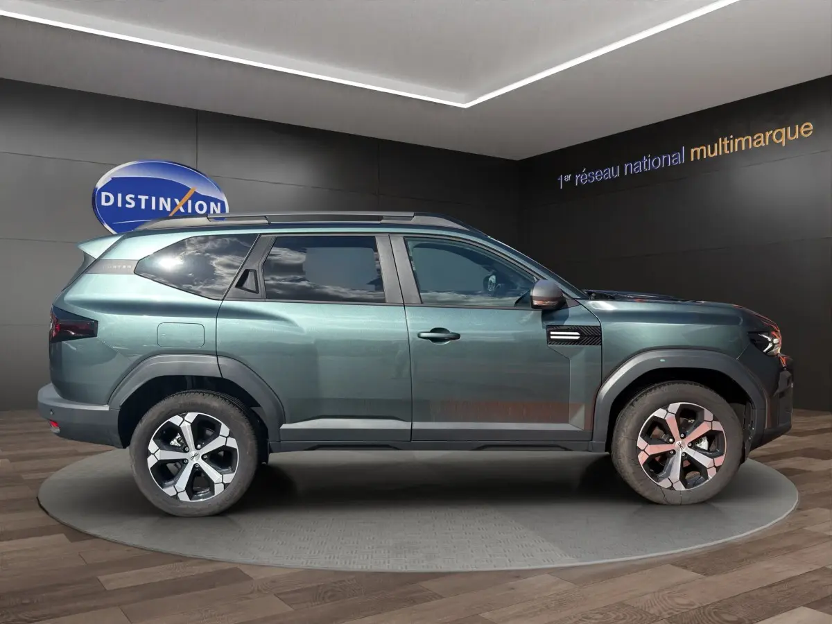 Vue de profil côté gauche du SUV Dacia Bigster vert métallisé avec attelage RDSO, roues alliage et barres de toit noires.
