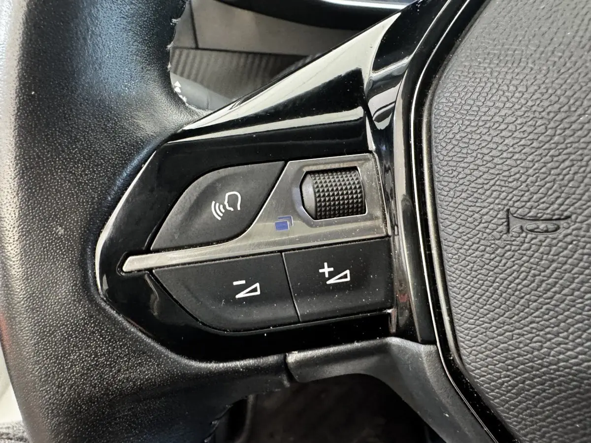Gros plan sur les commandes gauche du volant cuir noir du Peugeot 2008 blanc, avec boutons de volume et reconnaissance vocale.