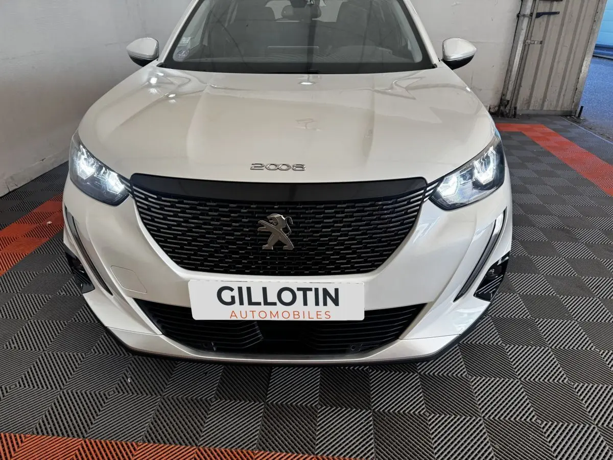 Vue de face d’un Peugeot 2008 blanc avec calandre noire et phares LED allumés dans un garage.