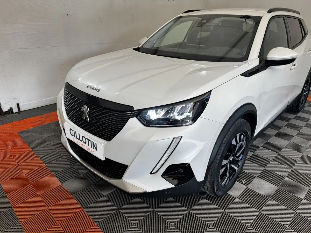Peugeot 2008 blanc en 3/4 avant droit, avec calandre noire et phares LED allumés sur sol à damiers.