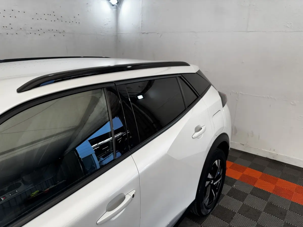 Vue 3/4 arrière droite d’un Peugeot 2008 blanc avec vitres teintées et barres de toit noires dans un garage.