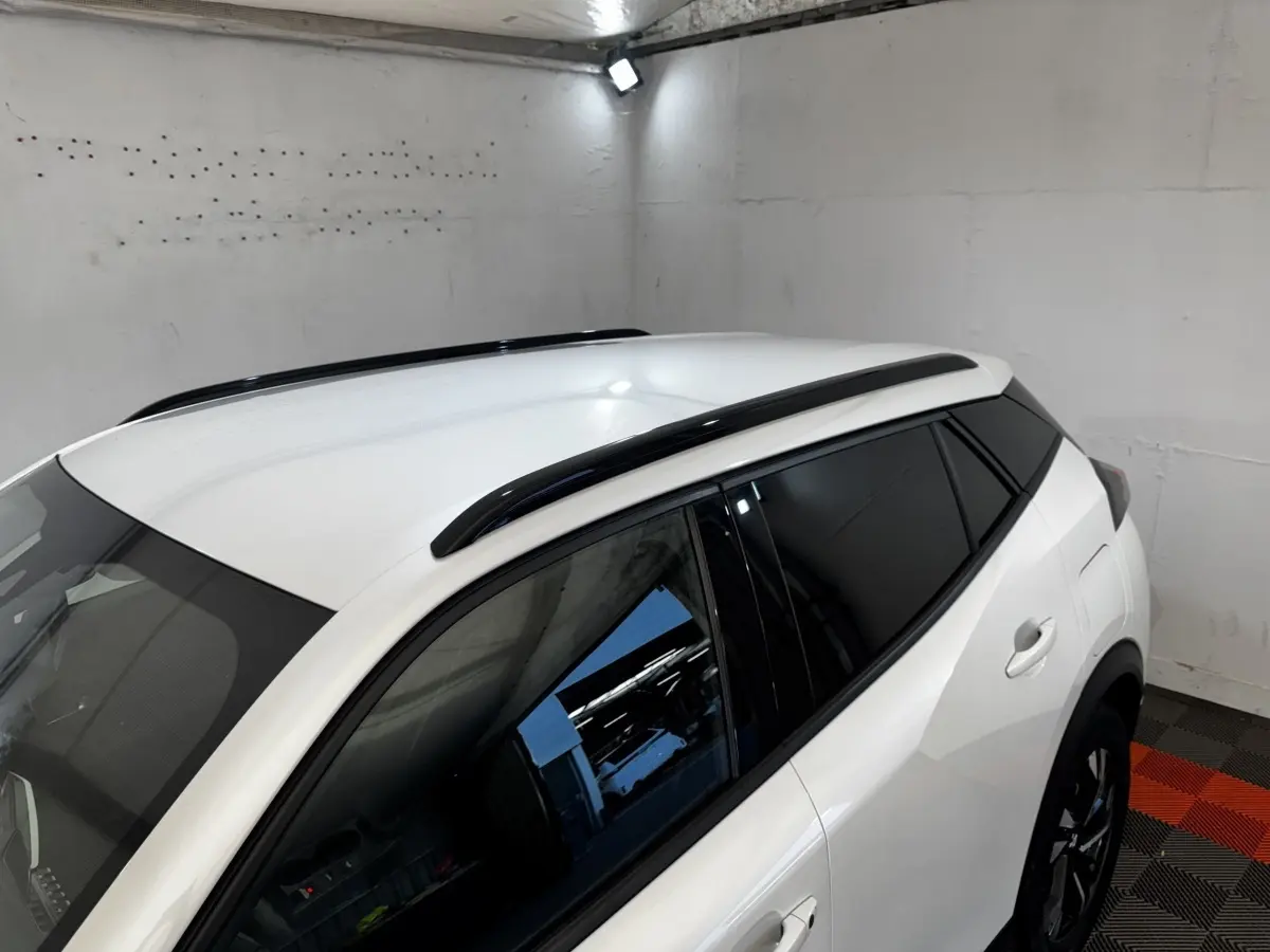 Vue 3/4 arrière droite du Peugeot 2008 blanc avec barres de toit noires et vitres teintées dans un garage.