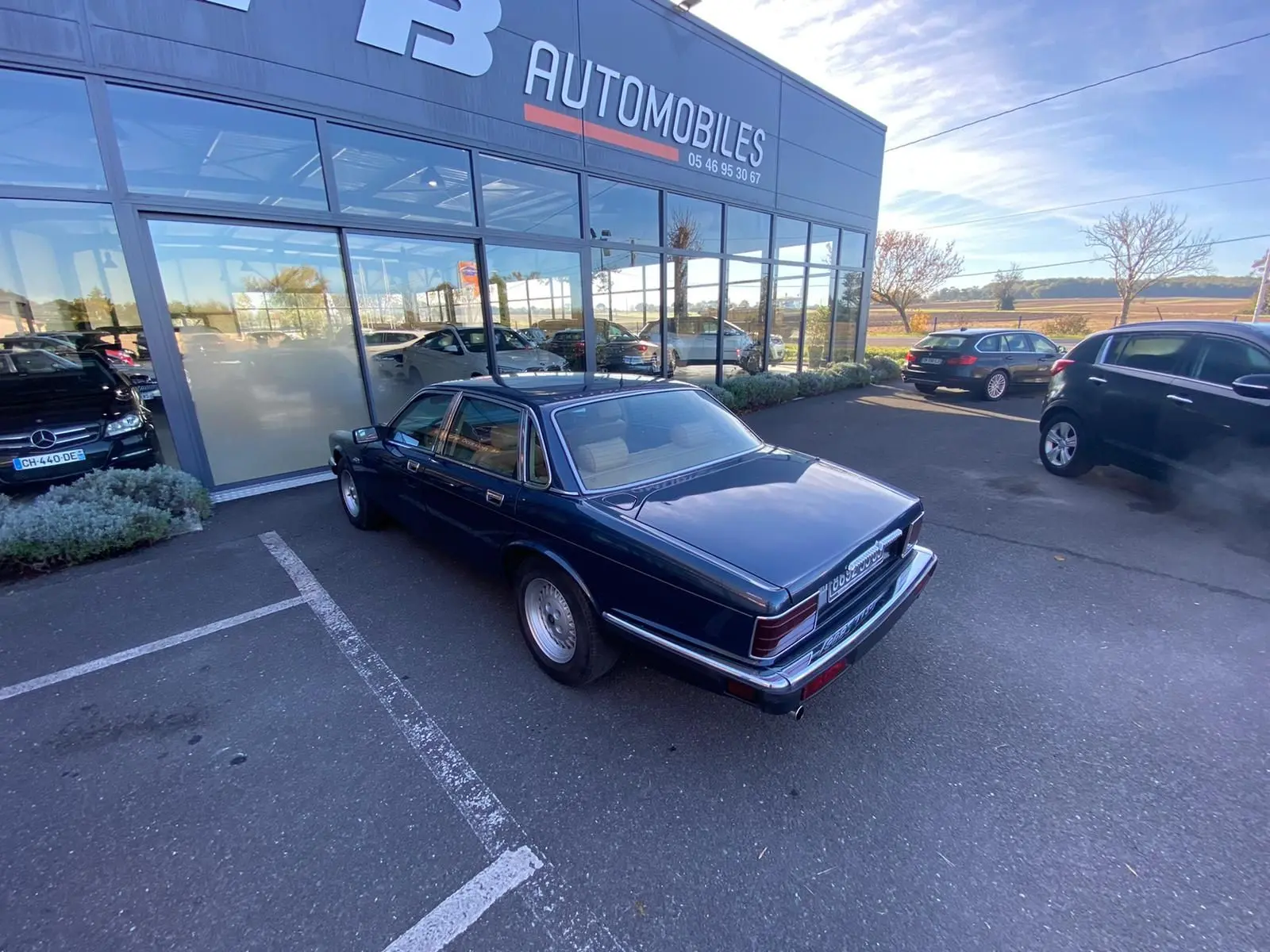 Jaguar XJ6 3.6 Sovereign bleu vue 3/4 arrière côté gauche, garée devant un showroom avec plusieurs autres voitures.