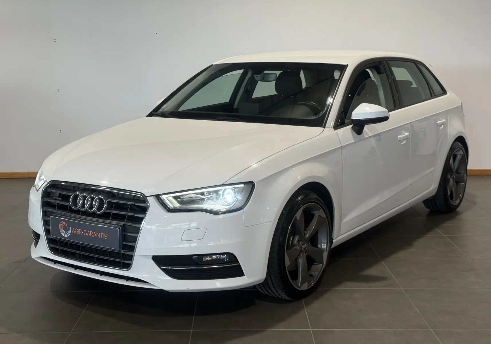 Audi A3 Sportback blanc vue 3/4 avant droit avec phares allumés et jantes foncées distinctives.