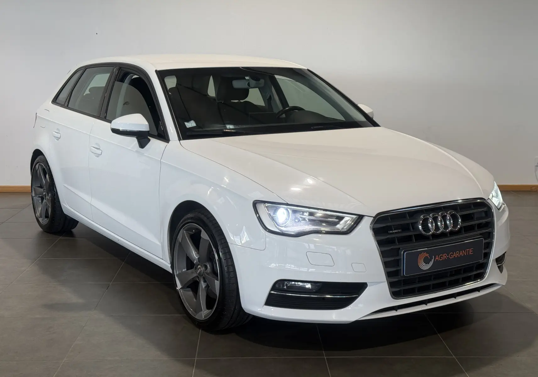 Audi A3 Sportback blanc vue 3/4 avant droit, phares allumés et jantes alliage foncé distinctives.