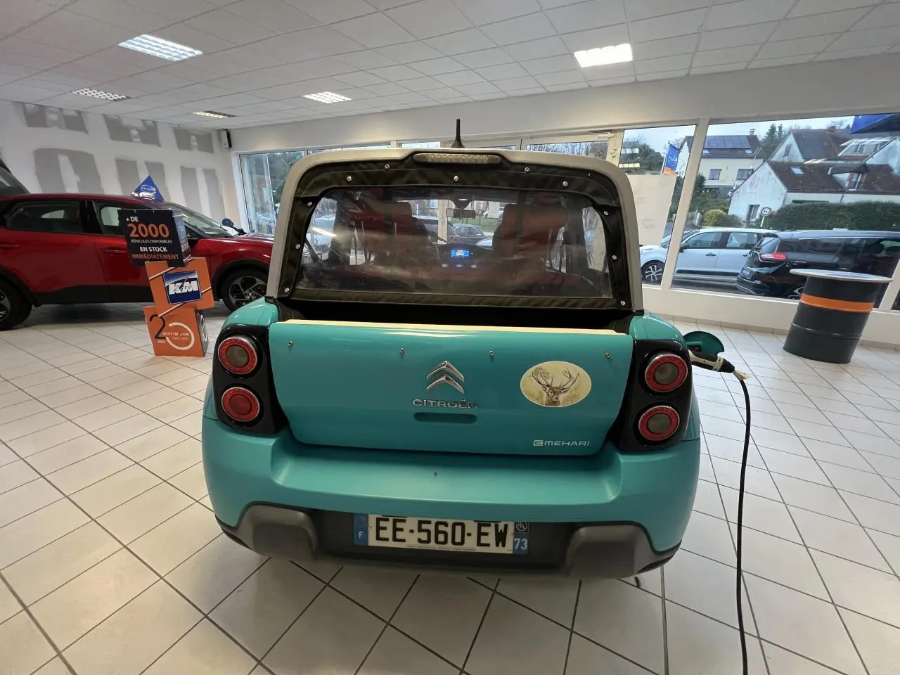 Vue arrière d'une Citroën E-MEHARI cabriolet bleu clair en recharge, avec toit en toile et autocollant cerf sur le hayon.
