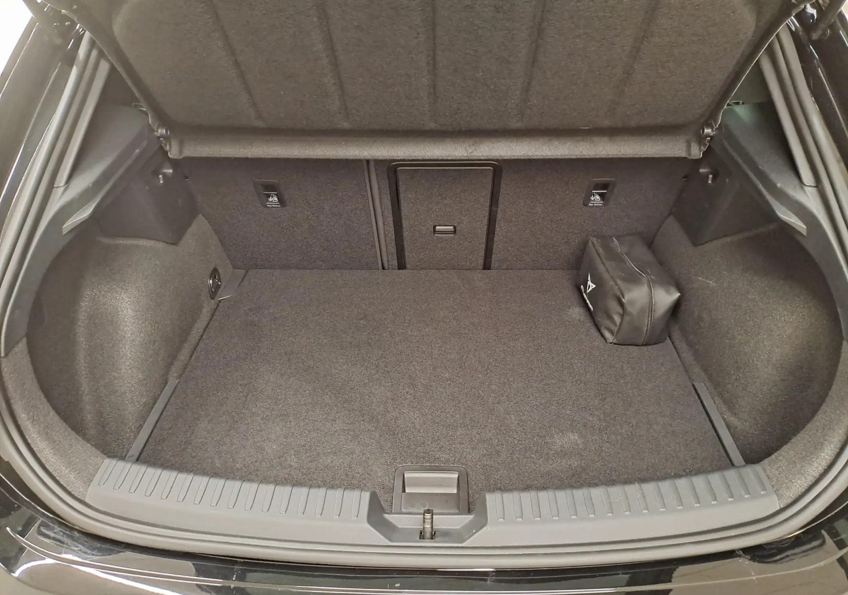 Coffre ouvert vu de l'arrière d'une CUPRA Leon Noir Minuit avec tapis gris et trousse de secours sur le côté droit.
