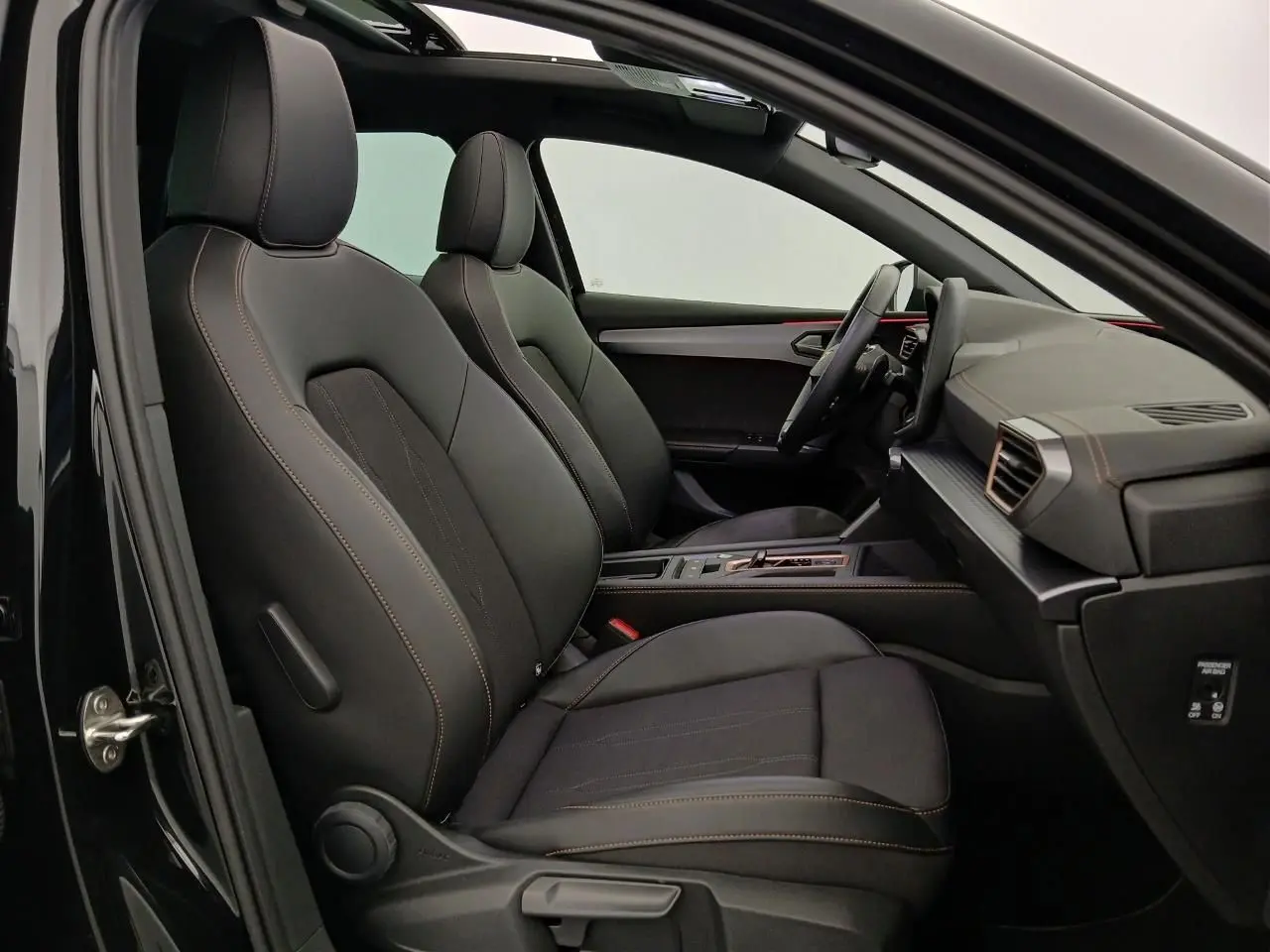 Vue intérieure côté gauche de la CUPRA Leon 2025 avec sièges noirs, surpiqûres orange et toit ouvrant panoramique visible.