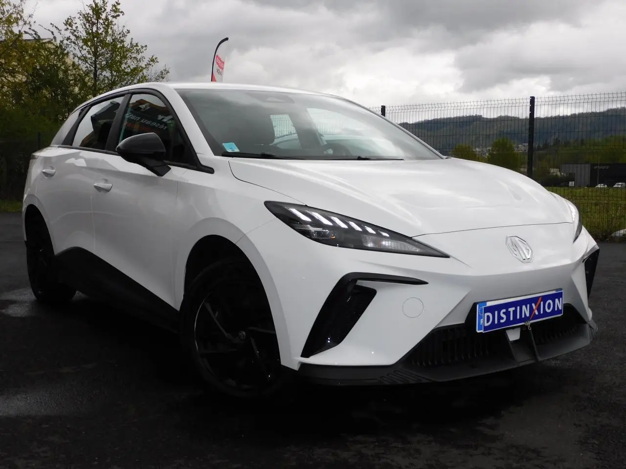 Vue 3/4 avant d'une MG4 électrique blanche avec jantes noires et feux LED distinctifs sur un parking extérieur.