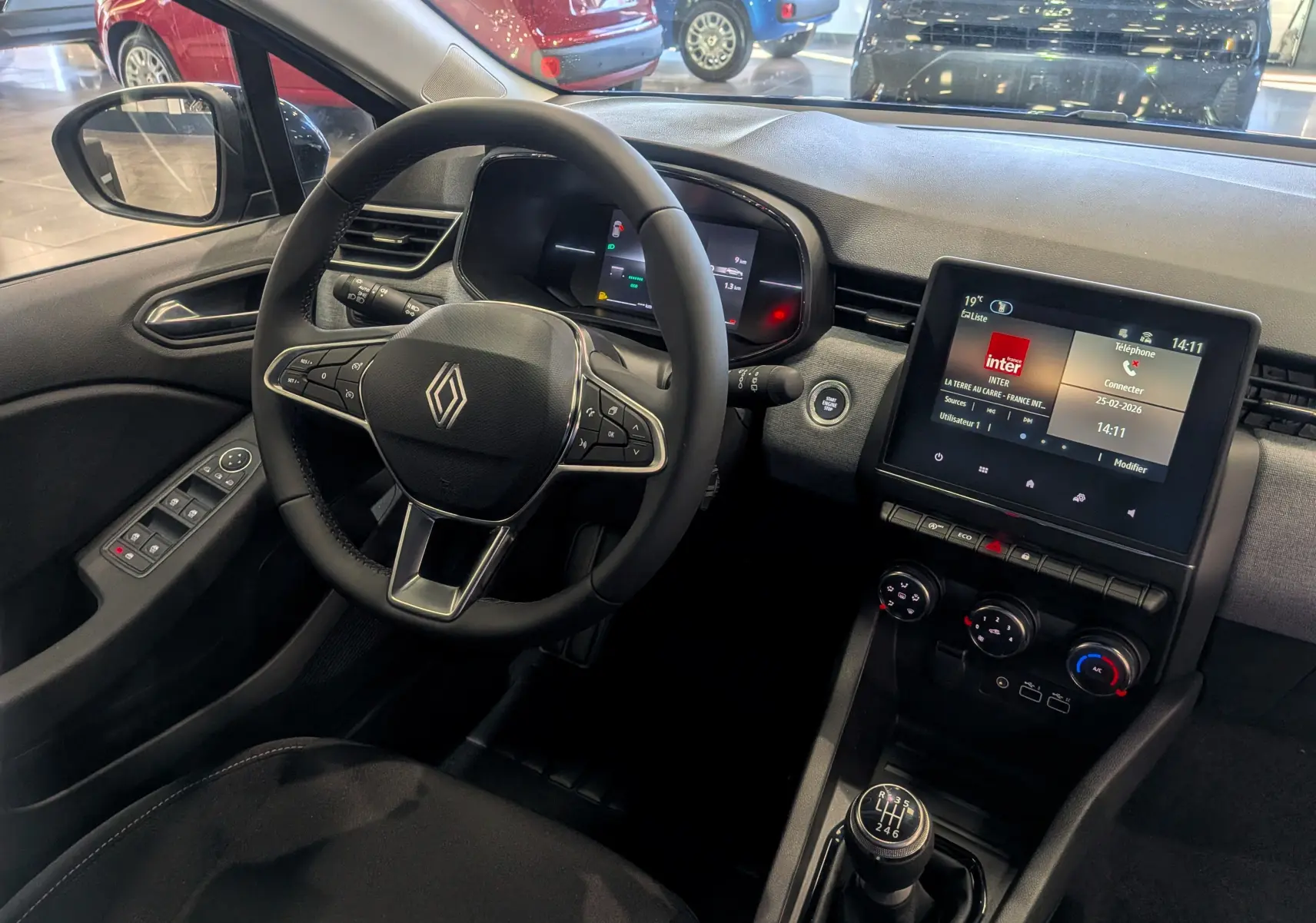 Vue intérieure côté conducteur de la Renault Clio V avec volant multifonction, écran tactile et levier de vitesse manuel.