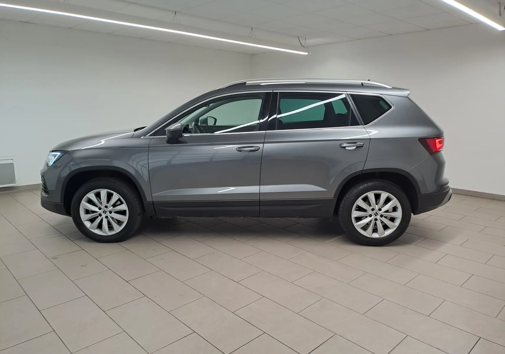Vue de profil côté gauche d'un SEAT Ateca 2023 gris graphite métallisé dans un intérieur lumineux.