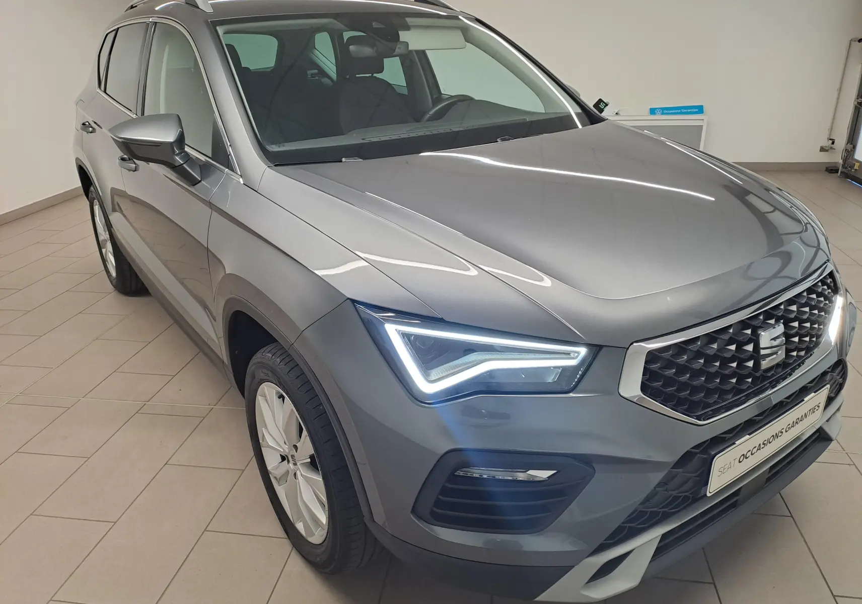 Vue 3/4 avant droit du SEAT Ateca 2023 gris graphite avec phares LED allumés et calandre distinctive en intérieur showroom.