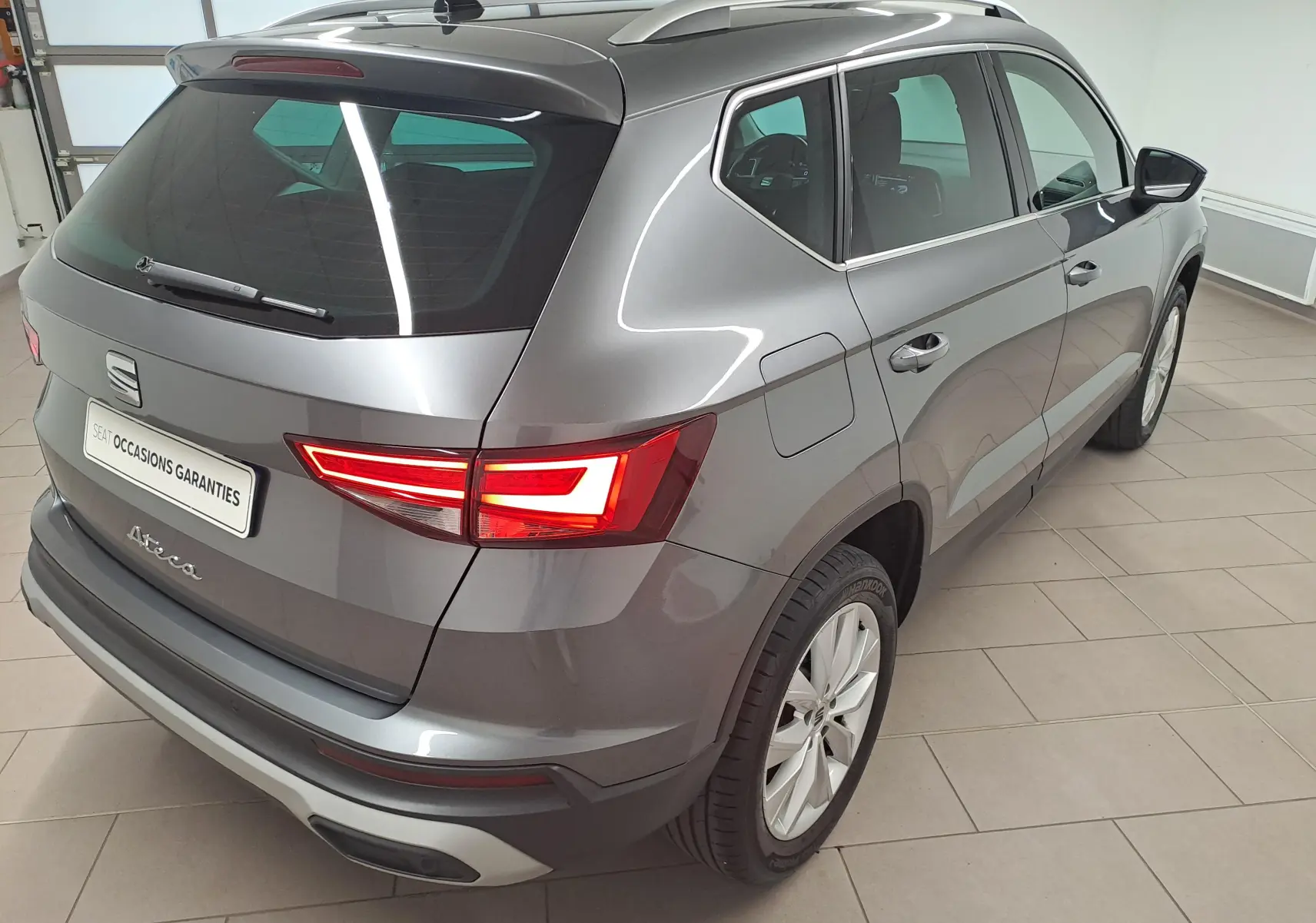 Vue 3/4 arrière droite d'un SEAT Ateca gris graphite métallisé, avec feux arrière allumés dans un showroom intérieur.