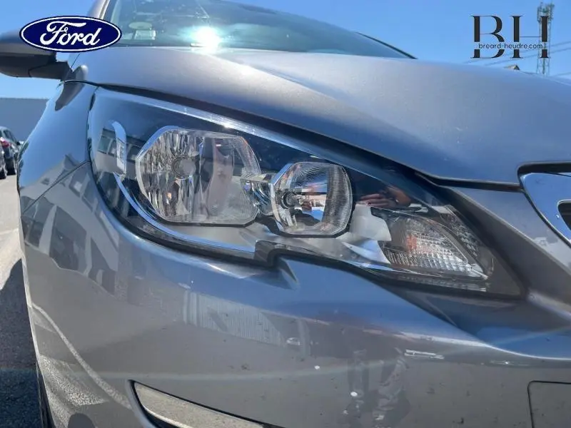Gros plan sur le phare avant droit d'une Peugeot 308 gris Artense sous un ciel ensoleillé.