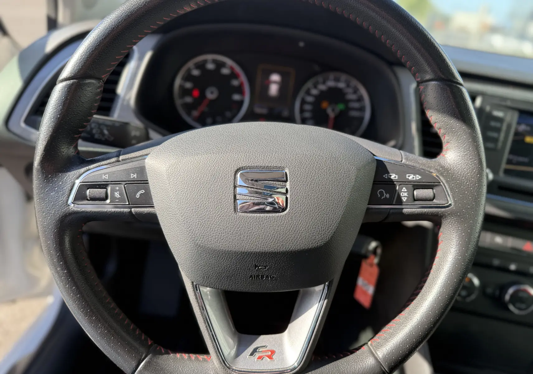 Vue rapprochée du volant noir avec surpiqûres rouges du SEAT LEON 1.8 TFSI 180 FR, mettant en valeur le logo FR et les commandes intégrées.