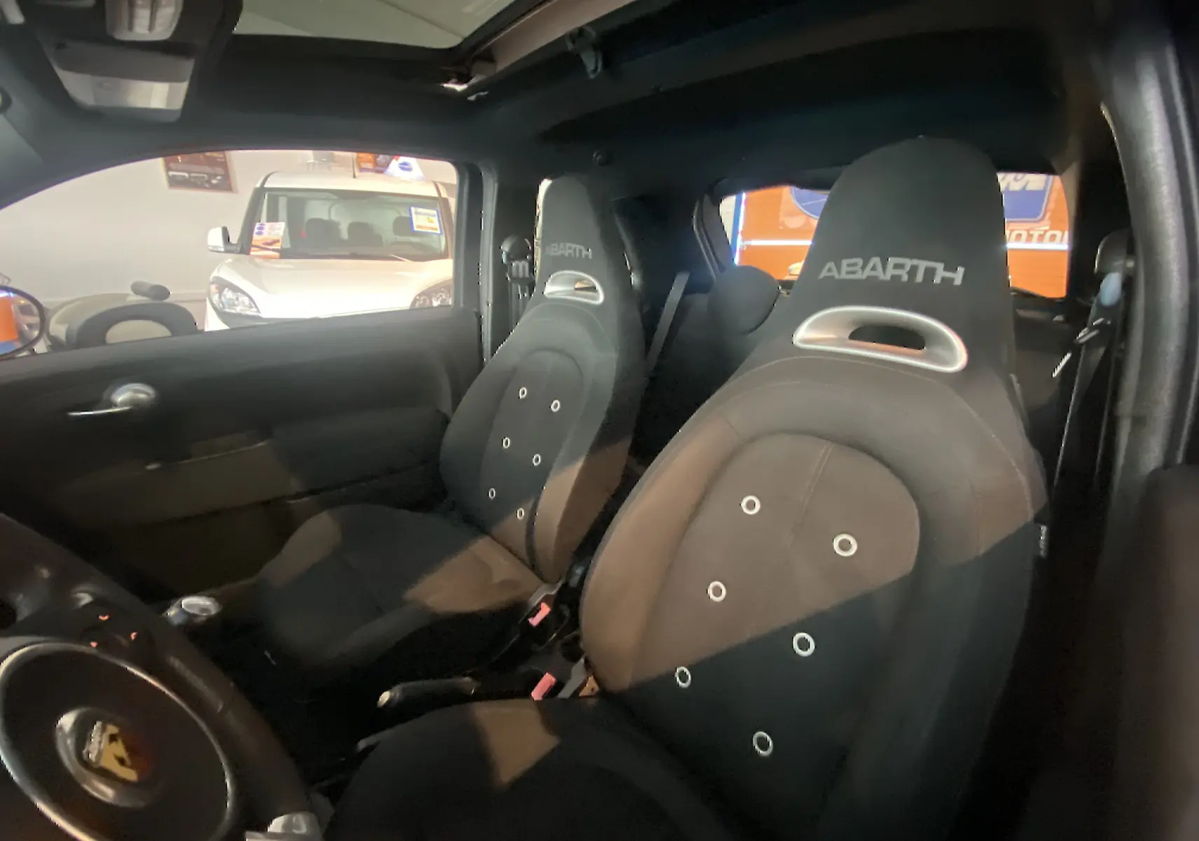 Vue intérieure côté conducteur de l'Abarth 595 noire, sièges sport avec logo ABARTH et toit ouvrant électrique visible.