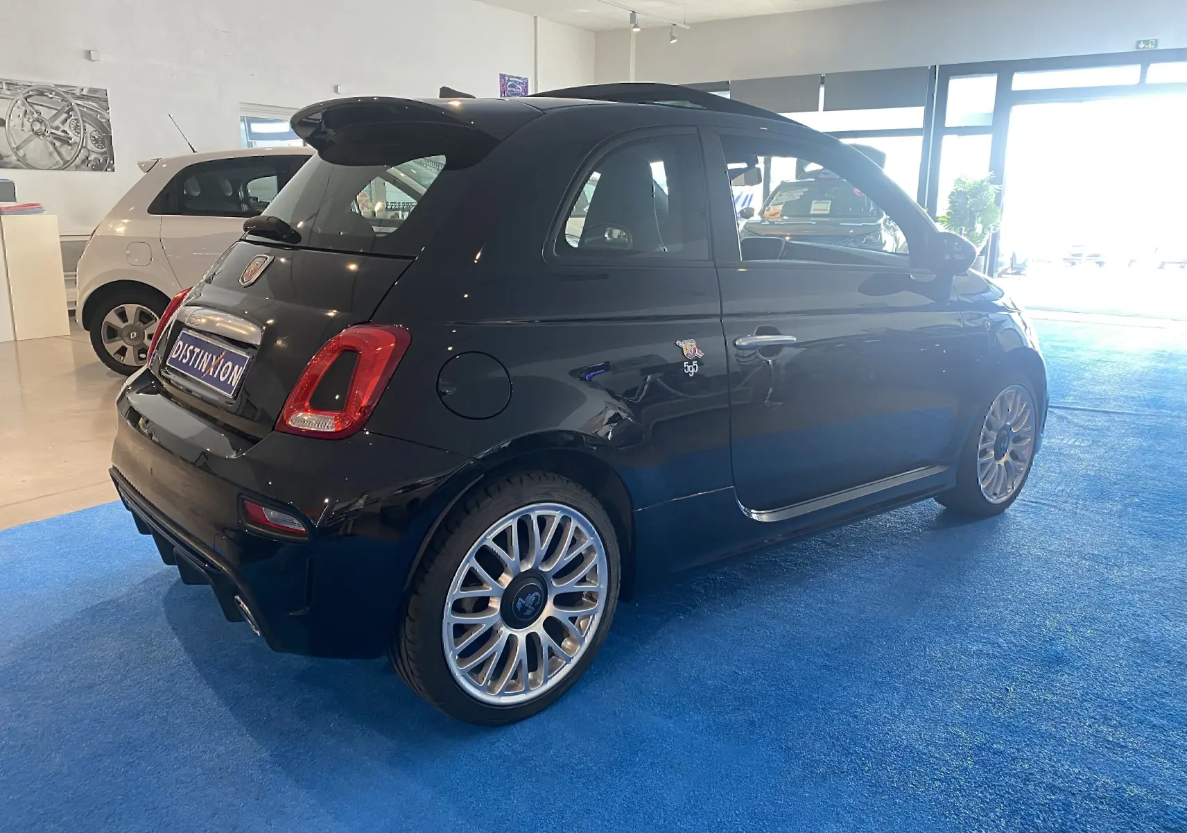 ABARTH 595 noir en vue 3/4 arrière droit, avec jantes alu 17 pouces et toit ouvrant électrique visible en intérieur showroom.