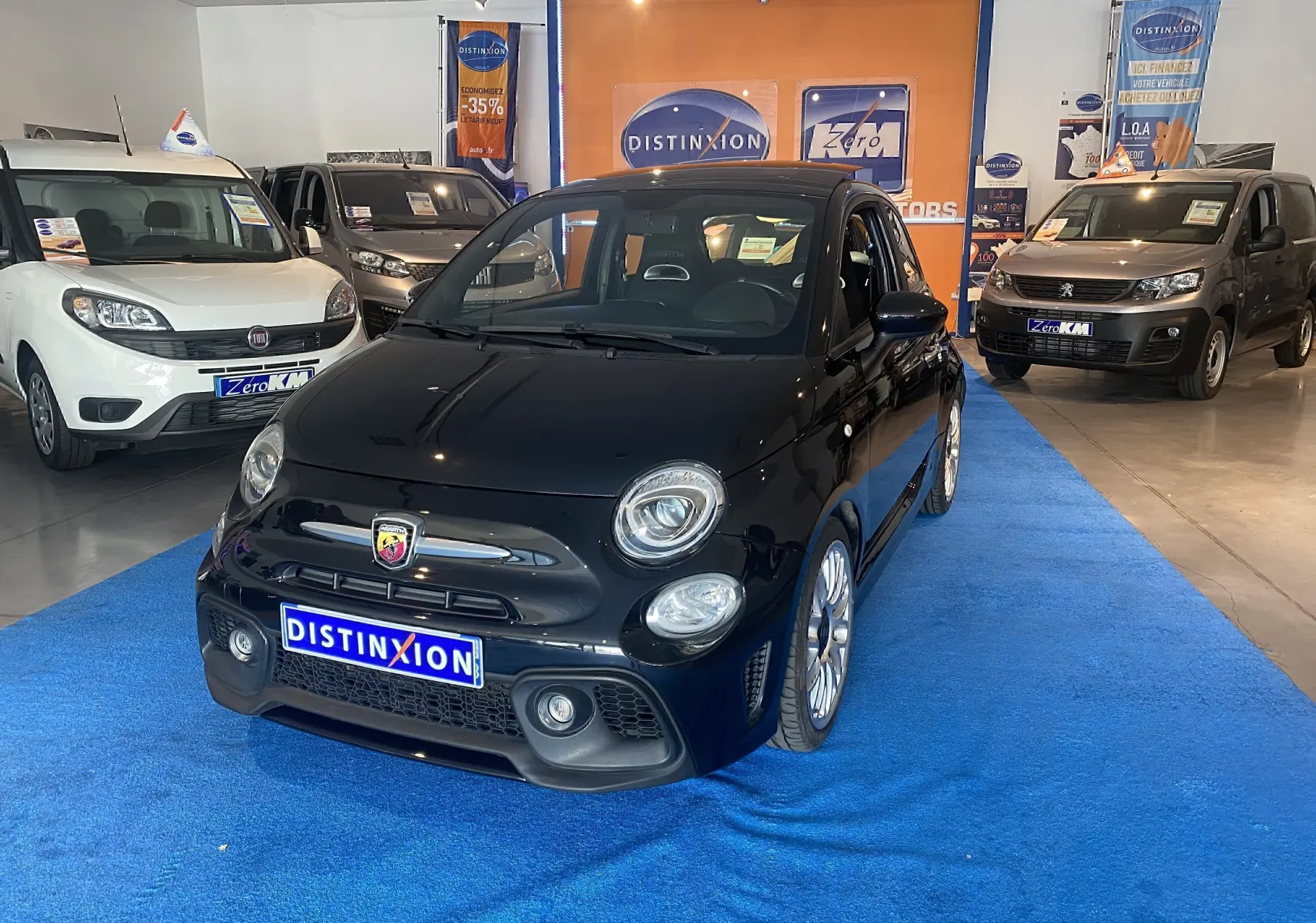 ABARTH 595 noir en 3/4 avant droit, jantes alu 17 pouces, toit ouvrant électrique, exposé en showroom sur tapis bleu.