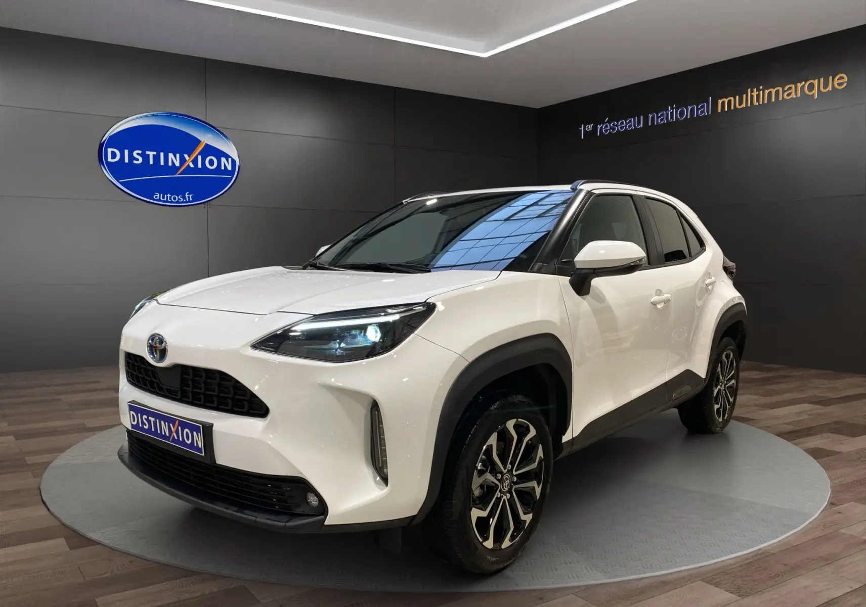 Toyota Yaris Cross blanc vue 3/4 avant droit, avec jantes alu 17 pouces et phares LED allumés dans showroom.