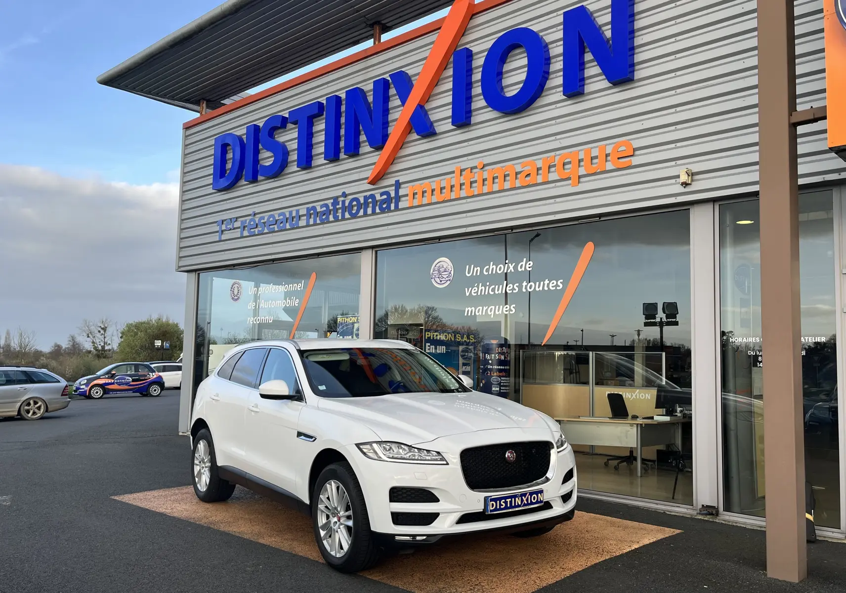 Jaguar F-PACE blanc en 3/4 avant droit, stationné devant un showroom Distinxion avec une calandre noire distinctive.